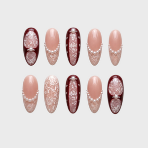 A set of press-on nails featuring burgundy frames, white lace patterns, and pearl embellishments on a nude base.