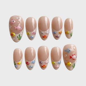 A set of nude French tip press-on nails decorated with 3D colorful fish, green seaweed, and clear water bubbles.
