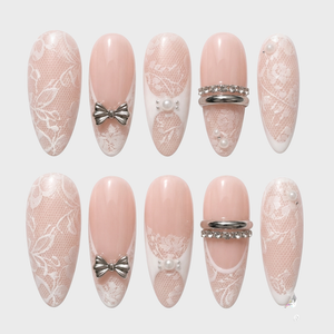 Stiletto press-on nails with nude base, white lace pattern, silver bows, and pearl accents.