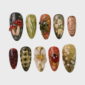A set of press-on nails featuring forest-themed 3D art including mushrooms, a frog, and a ladybug on green and terracotta backgrounds.