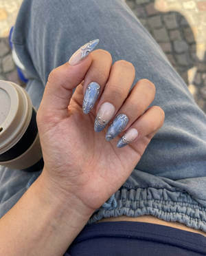 A set of press-on nails featuring a nude base, shimmering blue-grey French tips, and raised silver filigree swirl designs.