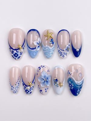 A set of ten press-on nails with blue and white floral patterns, gold starfish charms, pearls, and blue gems against a white background.
