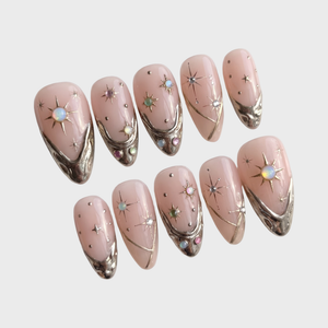 A set of pink press-on nails featuring gold metallic tips, celestial starburst details, and colorful iridescent gems.