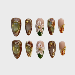 Brown press-on nails with 3D white floral designs, green gemstone centers, and gold metallic accents.