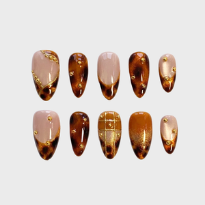 A set of tortoiseshell press-on nails featuring gold beads and gold leaf accents.