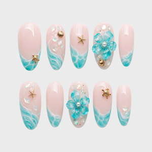 A set of sheer pink press-on nails featuring turquoise ocean wave designs, gold sea turtle and starfish charms, and 3D blue flowers with pearl details.