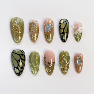 A set of press-on nails featuring olive green and pink bases decorated with gold butterfly wings, 3D flowers, and a large gemstone.