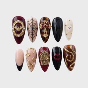 Gothic press-on nails featuring gold baroque embellishments, a bat, a cross, spiderwebs, and vampire lips on burgundy and black bases.