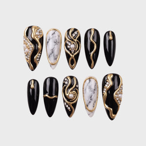 Black press-on nails with white marble patterns, gold swirls, pearls, and rhinestones.
