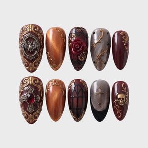 A set of gothic-themed press-on nails featuring burgundy, orange, and gray colors with gold bat, skull, rose, and cross embellishments.