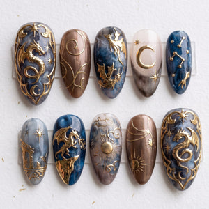 A set of press-on nails featuring detailed metallic gold dragons, celestial sun and moon motifs, and a blue and grey marble design.