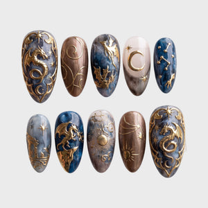 A set of press-on nails featuring detailed metallic gold dragons, celestial sun and moon motifs, and a blue and grey marble design.