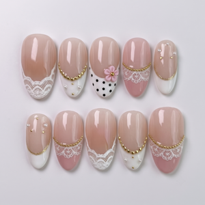 A set of press-on nails with white lace patterns, black polka dots, 3D pink flowers, gold beads, and pearl embellishments.