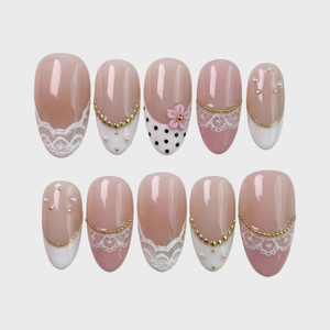 A set of press-on nails with white lace patterns, black polka dots, 3D pink flowers, gold beads, and pearl embellishments.