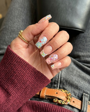 A set of press-on nails featuring a French tip design with floral motifs, gold foil, and glitter on a sheer base.
