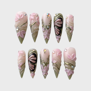 Stiletto press-on nails featuring 3D pink flowers, butterfly wings, sage green swirls, and rhinestones.