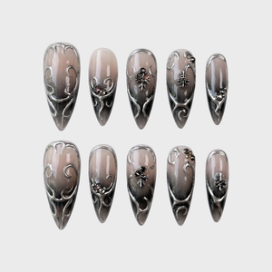 A set of stiletto press-on nails with a dark grey ombre gradient and silver filigree embellishments.