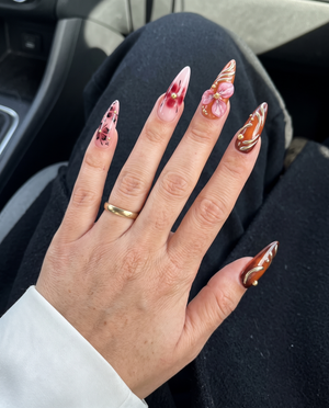 Stiletto shaped press-on nails featuring burgundy and nude polish with 3D floral accents, gold rose charms, and metallic swirl designs.