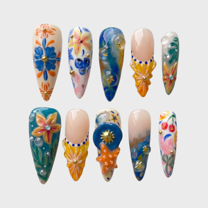 A set of ten stiletto-shaped press-on nails featuring a Mediterranean summer theme with 3D starfish, sun charms, yellow flowers, blue tile patterns, and ocean wave designs.