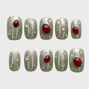 Sage green press-on nails featuring red ladybug motifs and clear dewdrop accents.