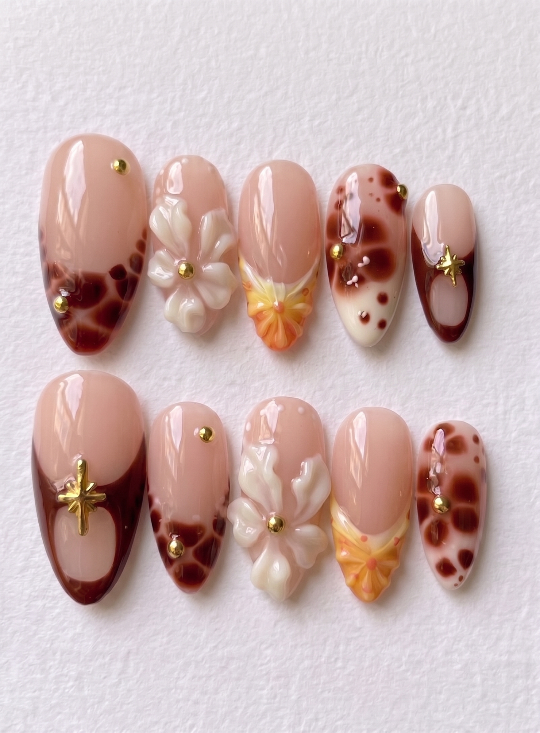 A set of press-on nails featuring tortoiseshell patterns, 3D white flowers, gold star charms, and yellow gradient details on a nude base.