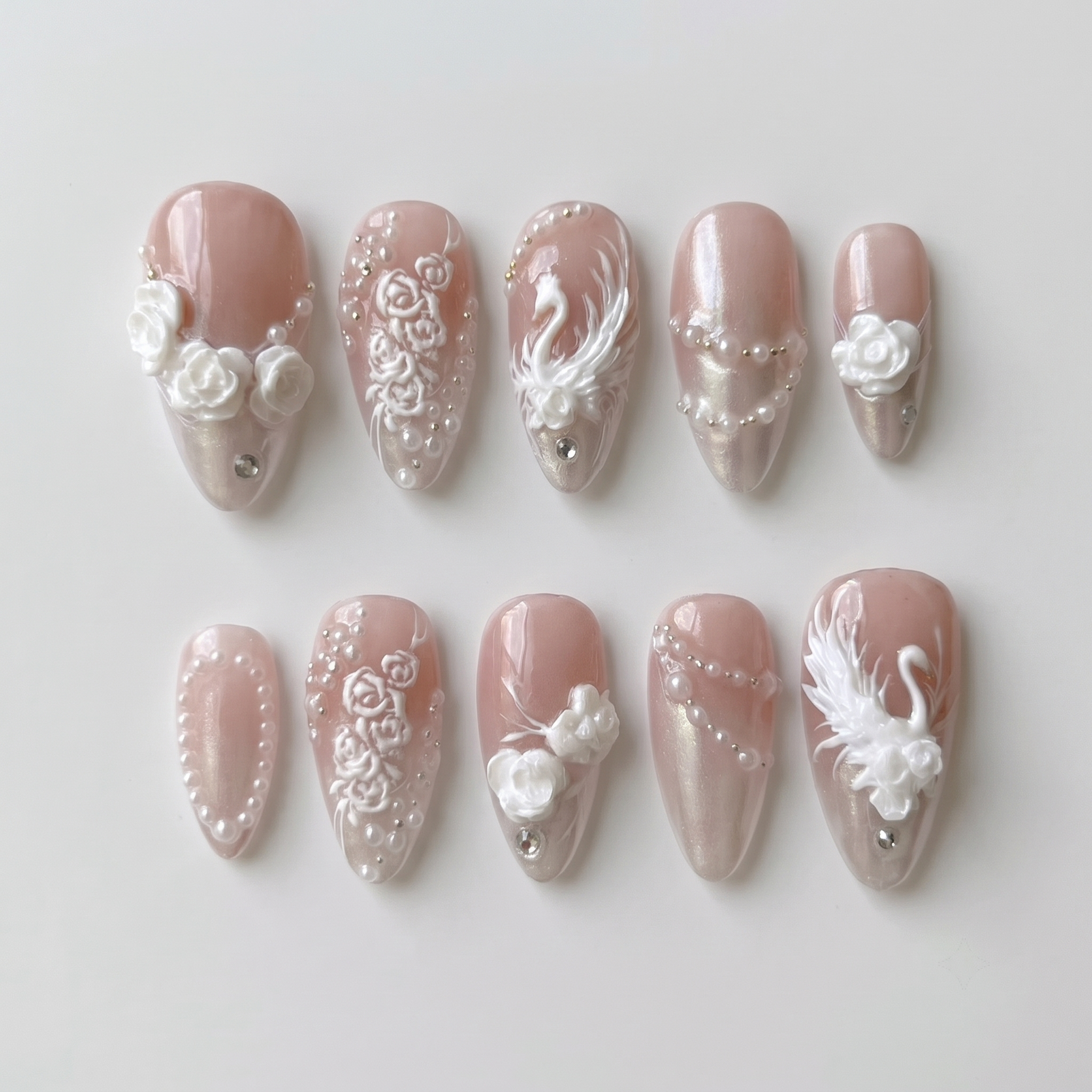 A set of press-on nails with 3D white roses, swan motifs, pearls, and rhinestones on a pearlescent nude base.
