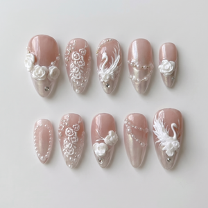 A set of press-on nails with 3D white roses, swan motifs, pearls, and rhinestones on a pearlescent nude base.