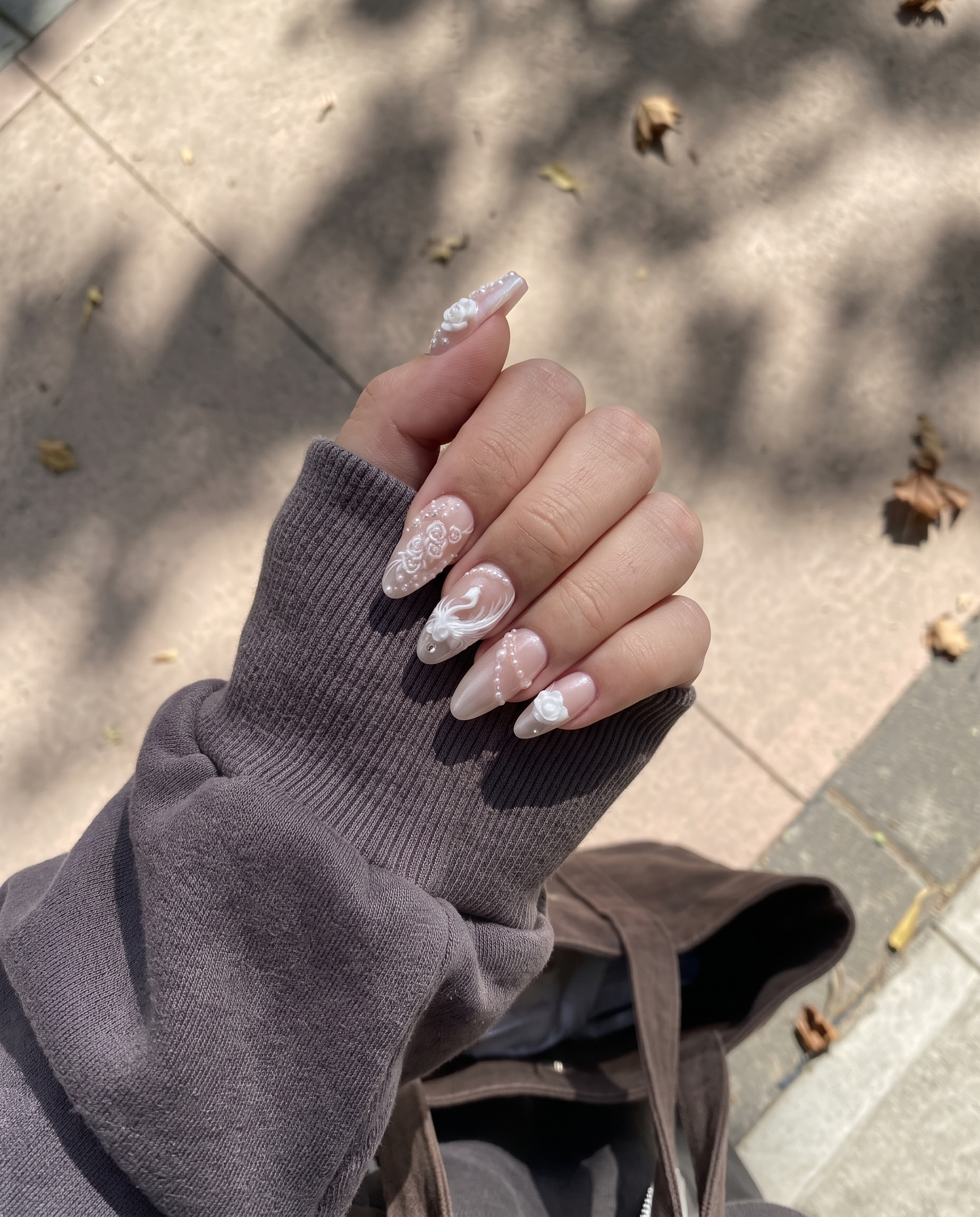 A set of press-on nails with 3D white roses, swan motifs, pearls, and rhinestones on a pearlescent nude base.