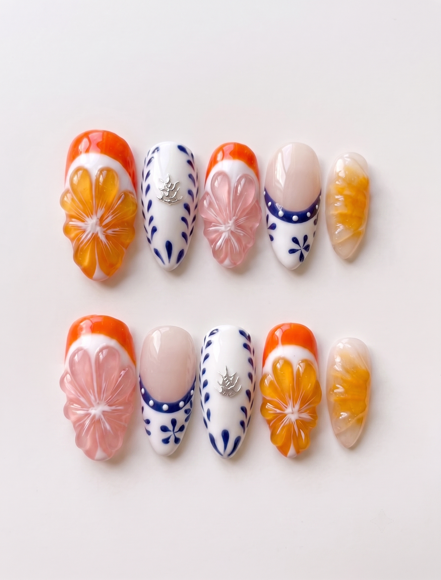 A set of press-on nails featuring 3D orange and pink fruit slices, blue floral patterns on white, and French tips.