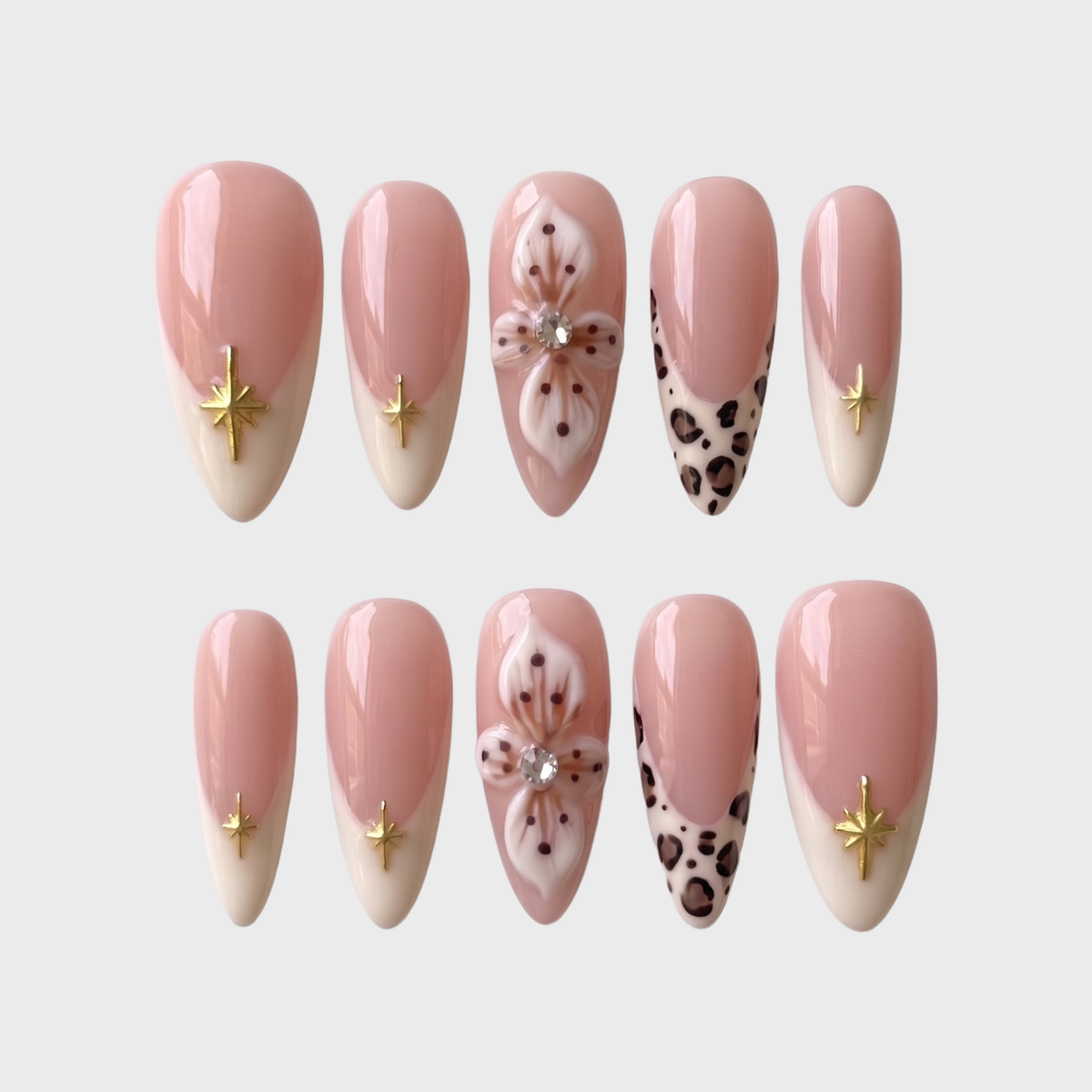 A set of nude pink press-on nails featuring cream French tips with gold stars, 3D white flowers with rhinestones, and leopard print tips.