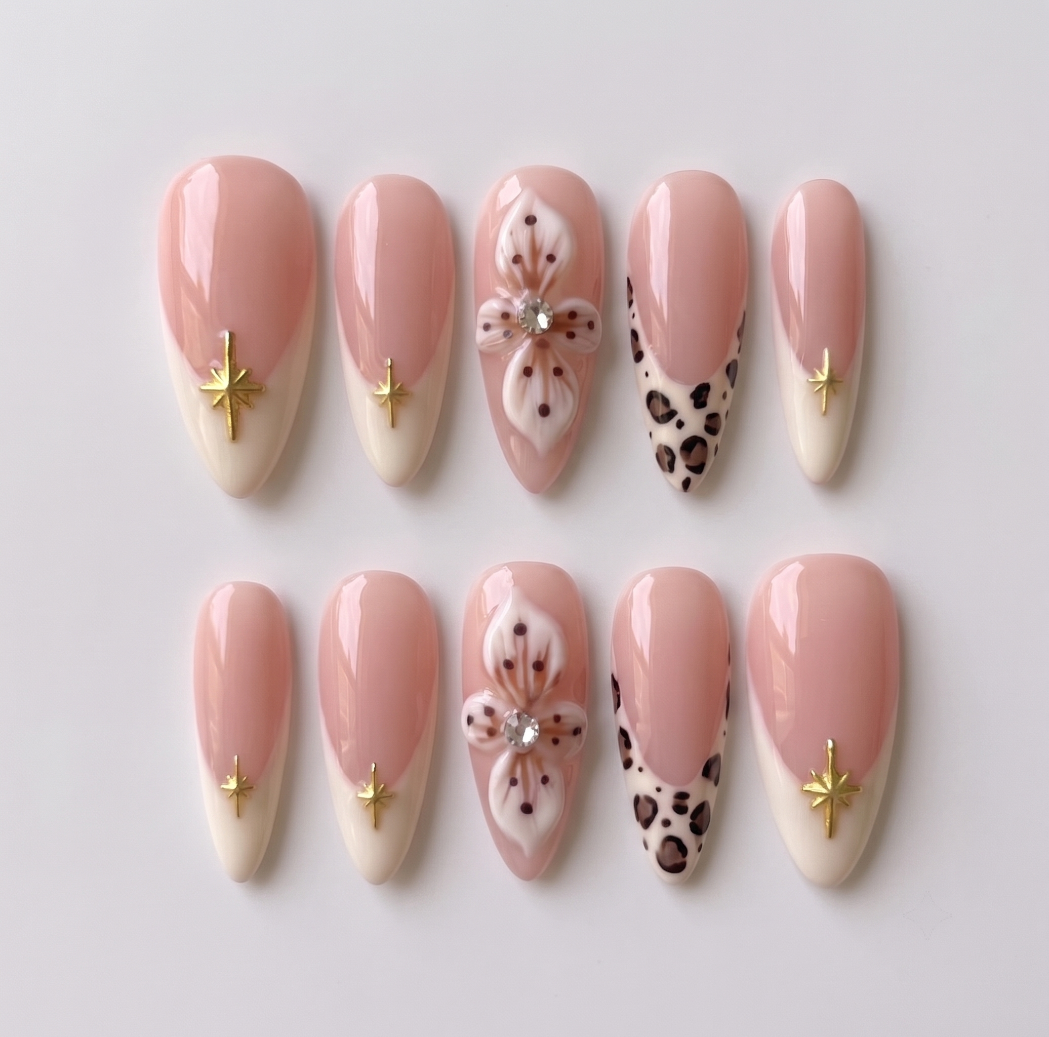 A set of nude pink press-on nails featuring cream French tips with gold stars, 3D white flowers with rhinestones, and leopard print tips.