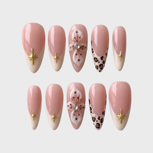 A set of nude pink press-on nails featuring cream French tips with gold stars, 3D white flowers with rhinestones, and leopard print tips.