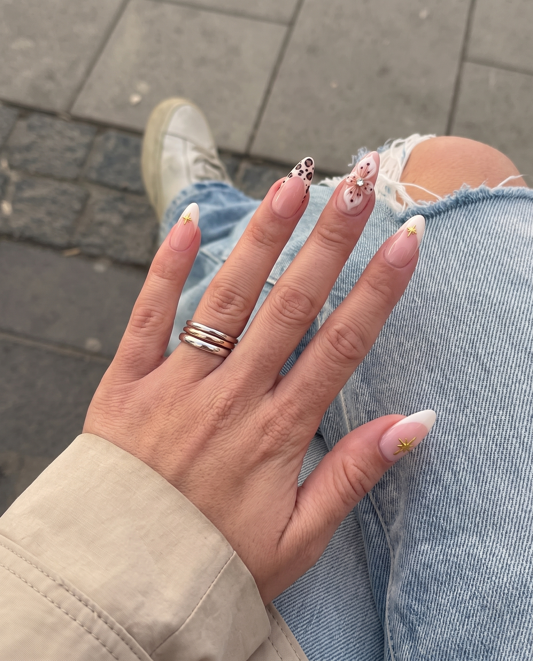 A set of nude pink press-on nails featuring cream French tips with gold stars, 3D white flowers with rhinestones, and leopard print tips.