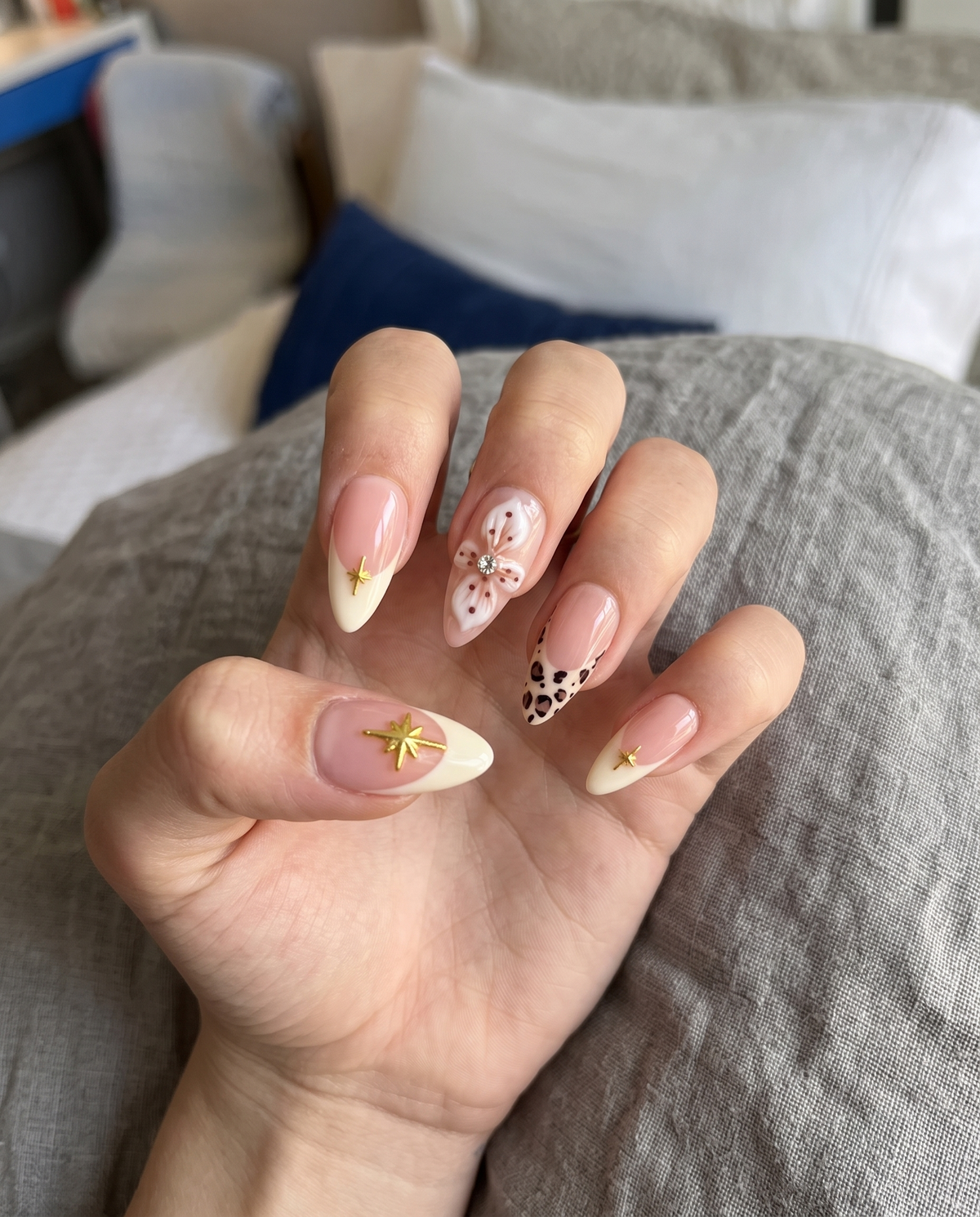 A set of nude pink press-on nails featuring cream French tips with gold stars, 3D white flowers with rhinestones, and leopard print tips.