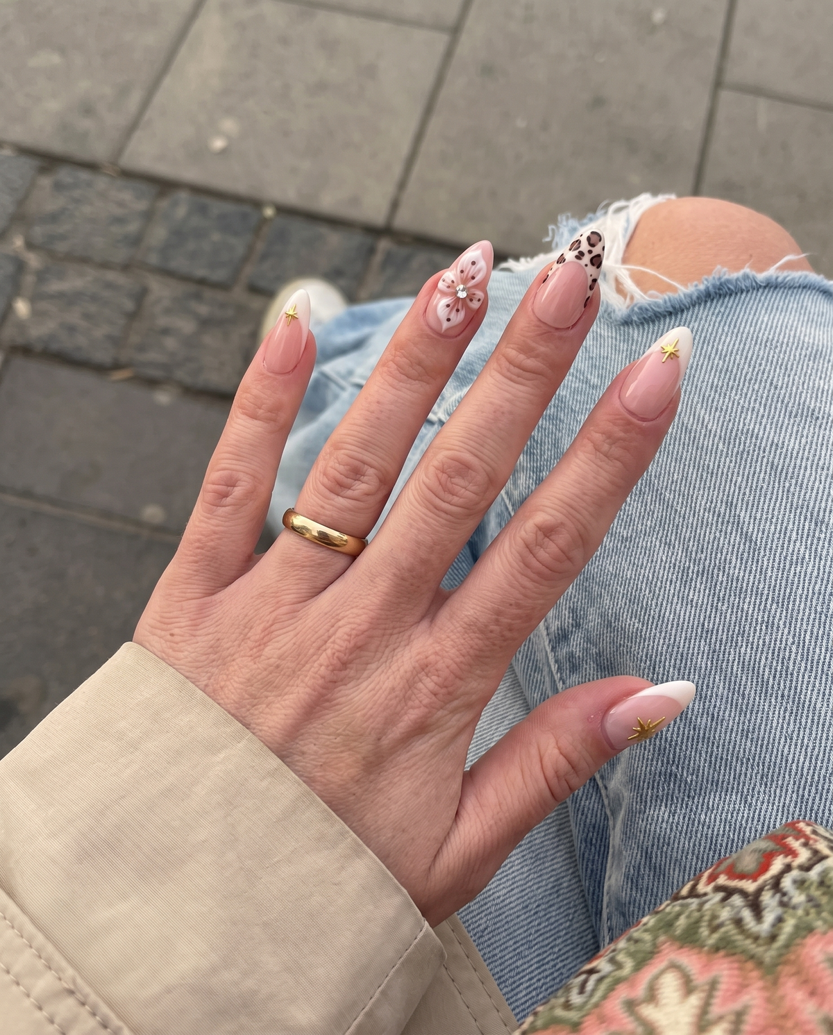 A set of nude pink press-on nails featuring cream French tips with gold stars, 3D white flowers with rhinestones, and leopard print tips.