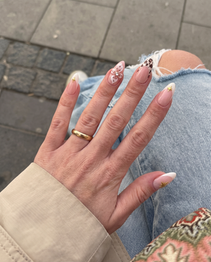 A set of nude pink press-on nails featuring cream French tips with gold stars, 3D white flowers with rhinestones, and leopard print tips.