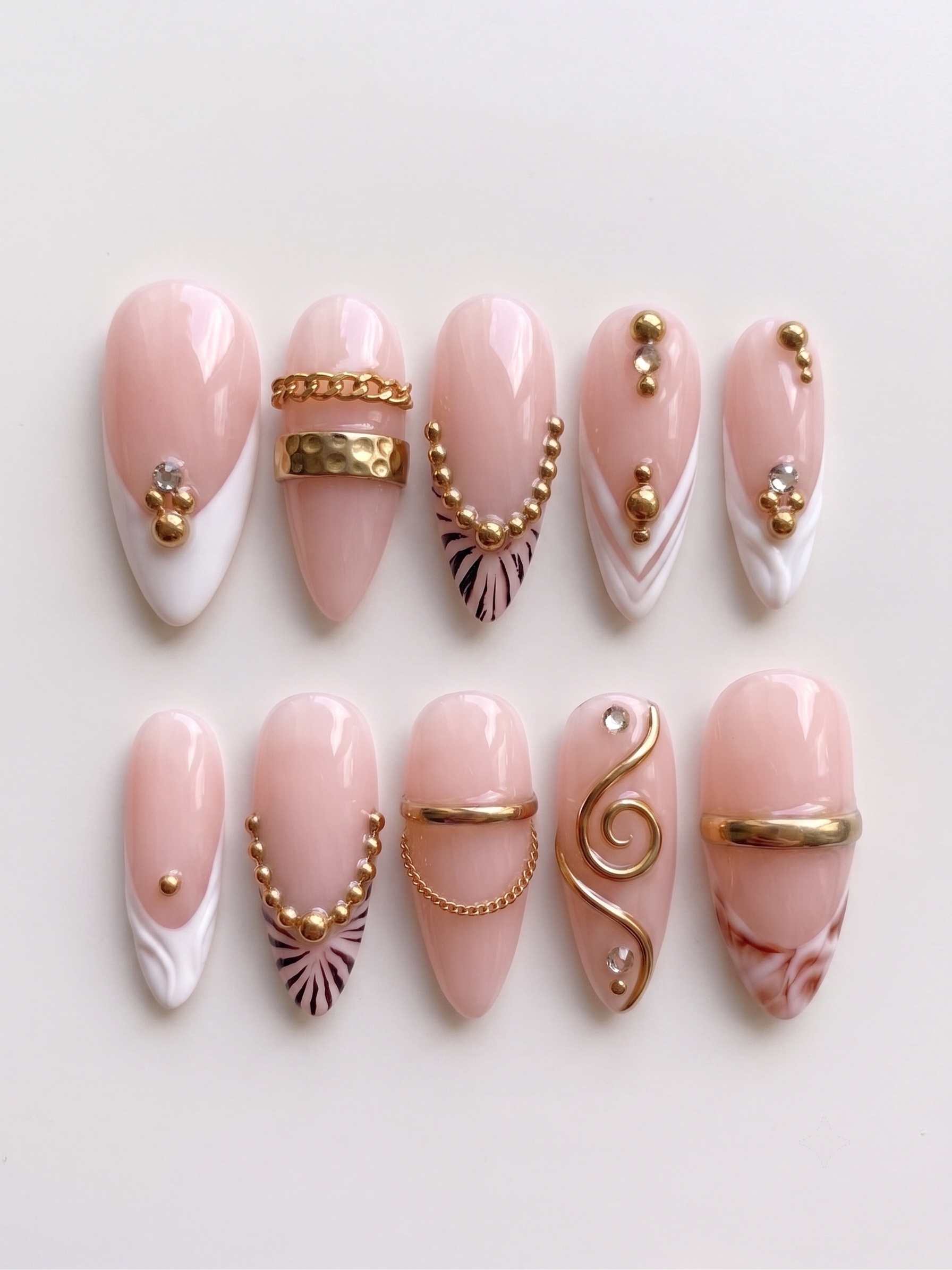 A set of pink and white press-on nails decorated with gold bands, chains, studs, zebra stripes, and rhinestones.