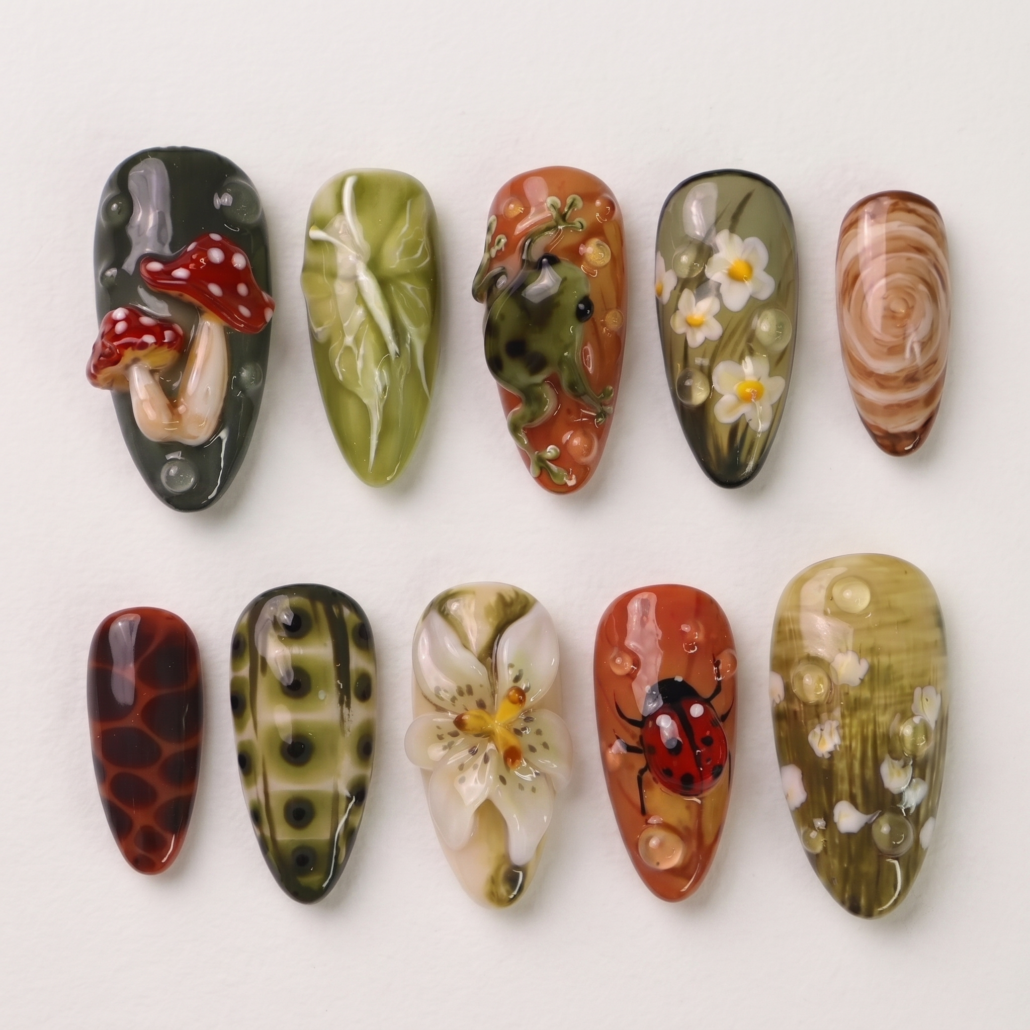 A set of press-on nails featuring forest-themed 3D art including mushrooms, a frog, and a ladybug on green and terracotta backgrounds.