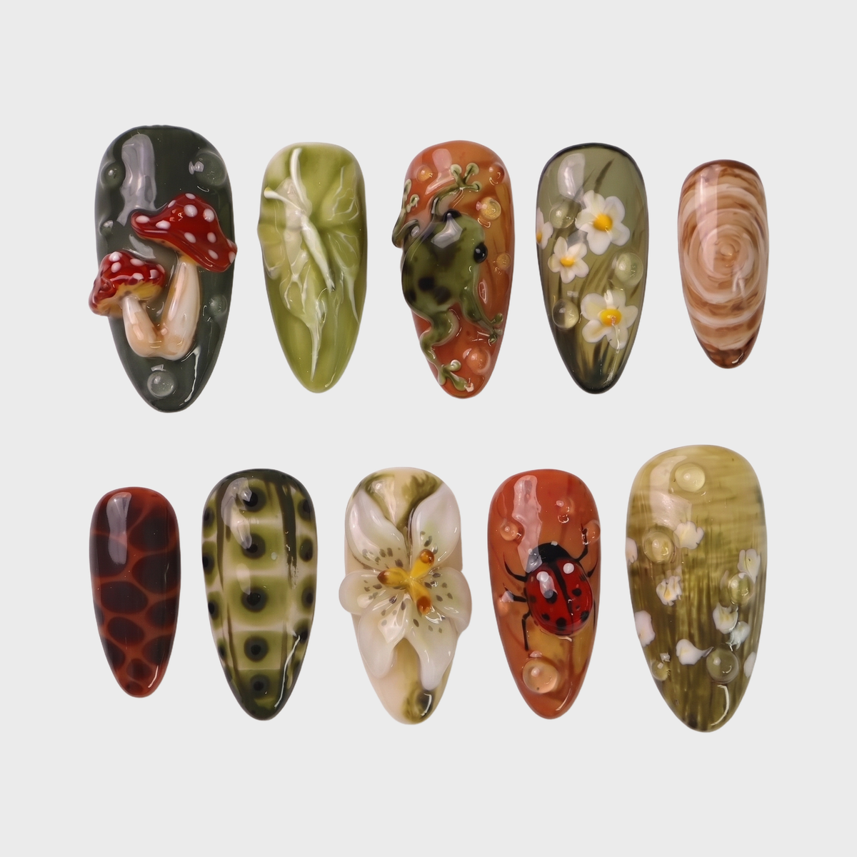 A set of press-on nails featuring forest-themed 3D art including mushrooms, a frog, and a ladybug on green and terracotta backgrounds.