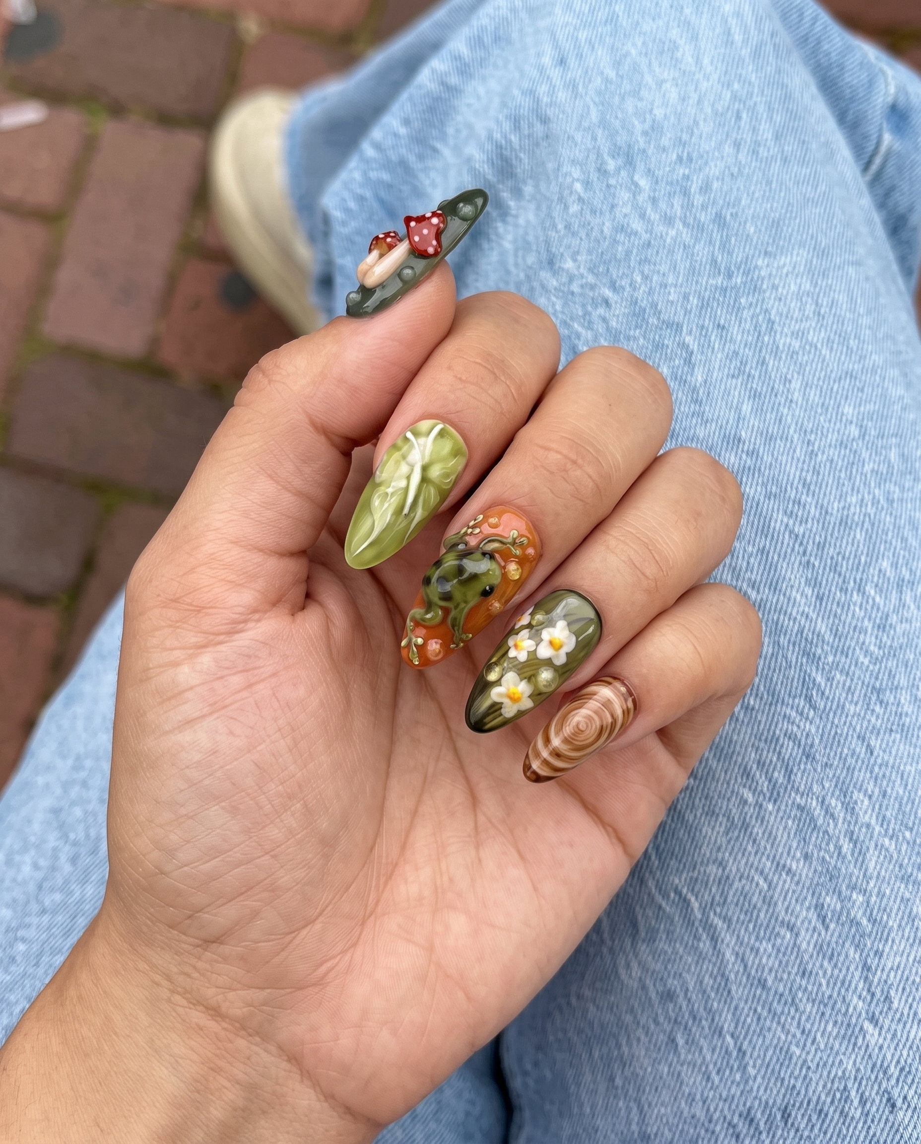 A set of press-on nails featuring forest-themed 3D art including mushrooms, a frog, and a ladybug on green and terracotta backgrounds.