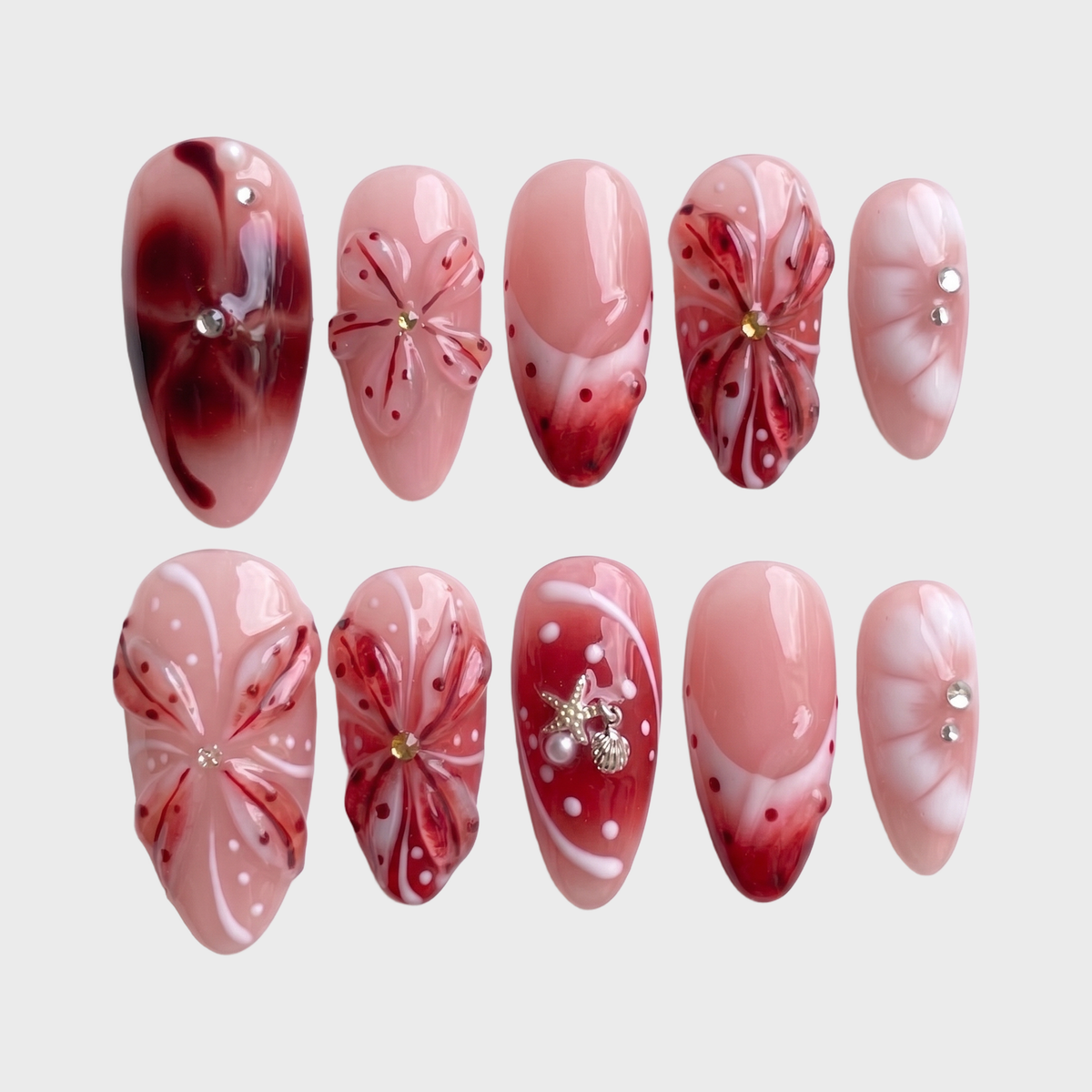 Set of press-on nails featuring red 3D floral designs, nautical charms, and rhinestone accents on a soft pink base.