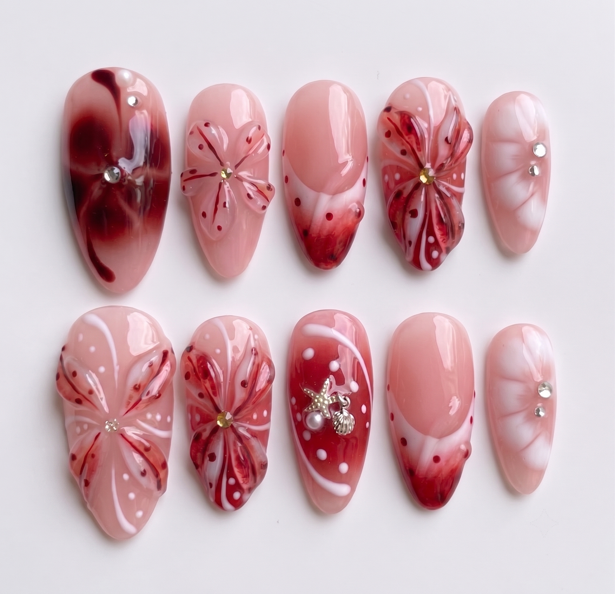 Set of press-on nails featuring red 3D floral designs, nautical charms, and rhinestone accents on a soft pink base.