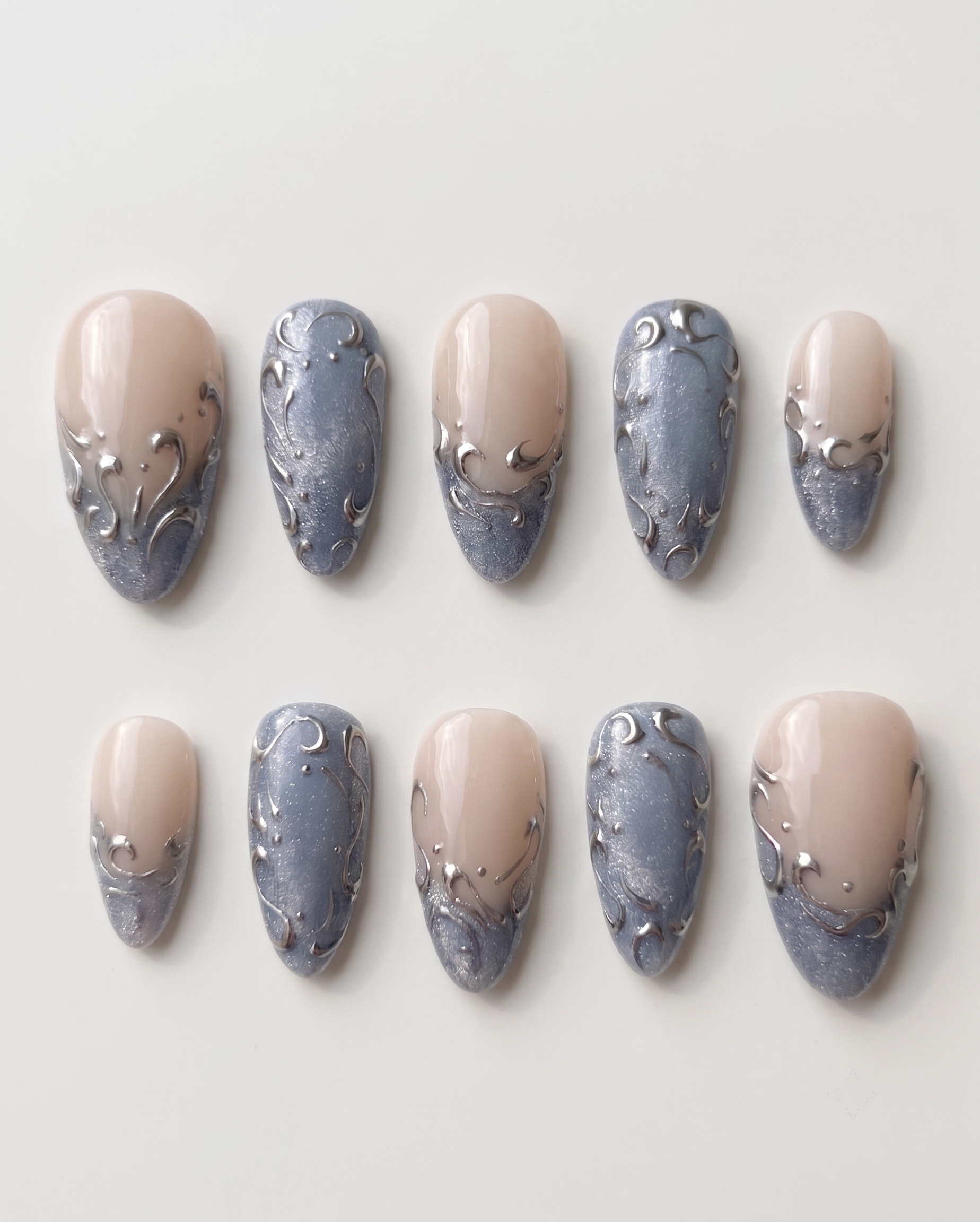 A set of press-on nails featuring a nude base, shimmering blue-grey French tips, and raised silver filigree swirl designs.