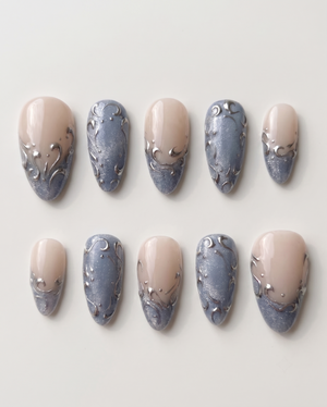 A set of press-on nails featuring a nude base, shimmering blue-grey French tips, and raised silver filigree swirl designs.
