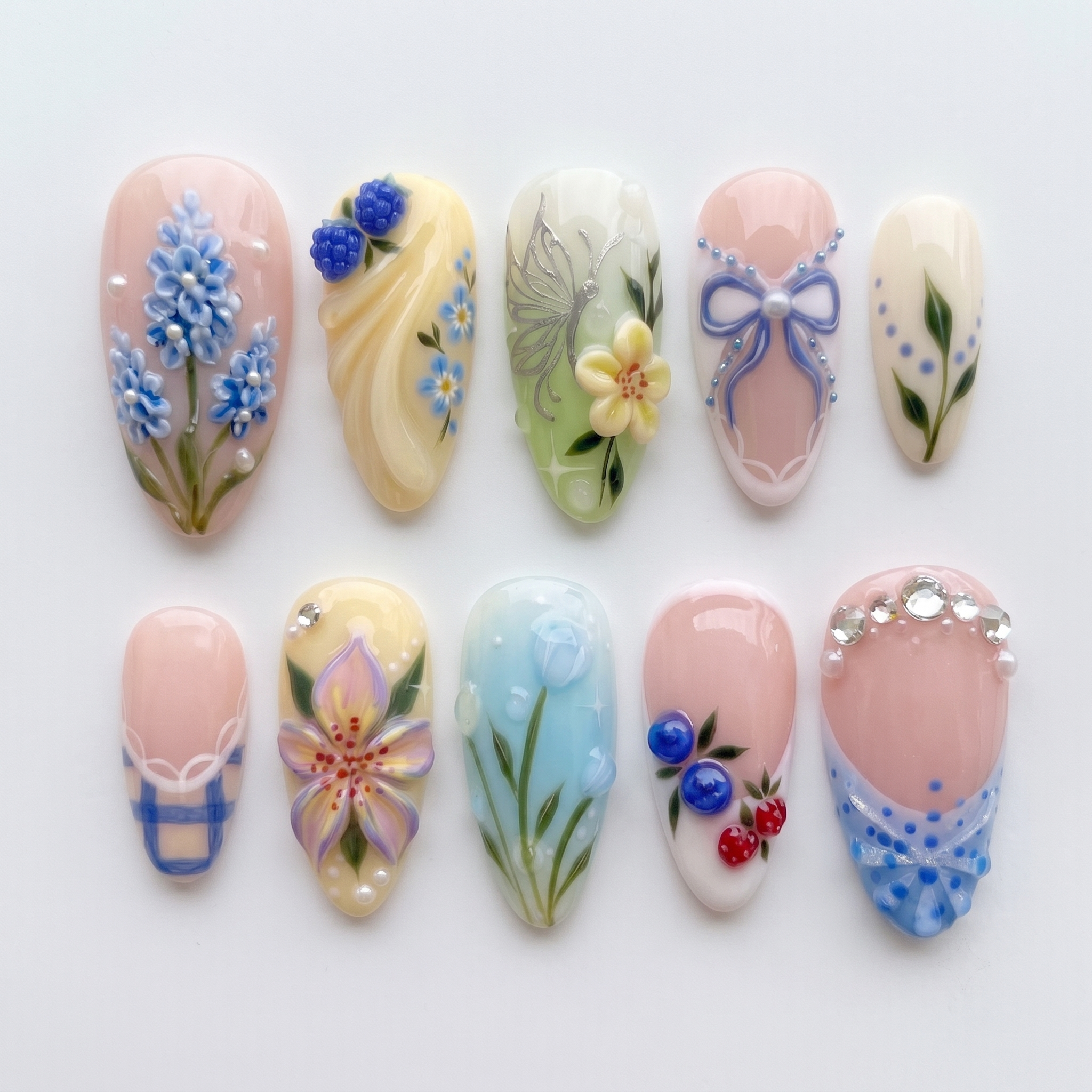 Mismatched pastel press-on nails featuring 3D blue flowers, berries, tulips, a silver butterfly, bows, and rhinestone embellishments.