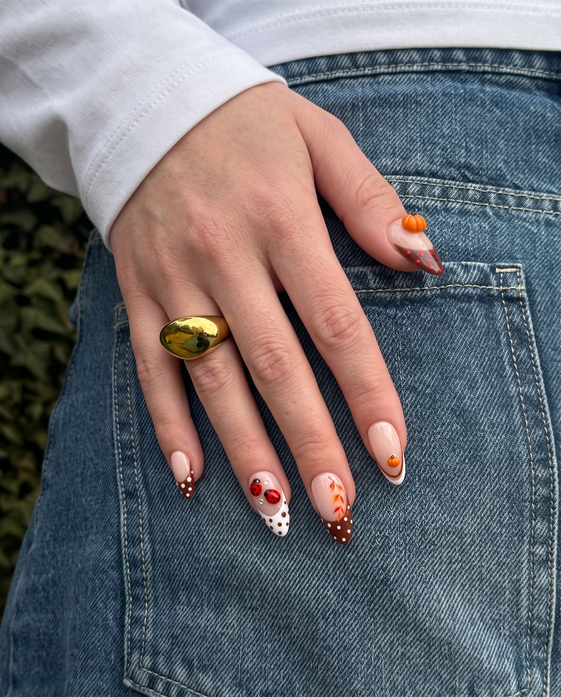 A set of press-on nails featuring brown French tips with white polka dots, decorated with 3D pumpkin charms and ladybug art.