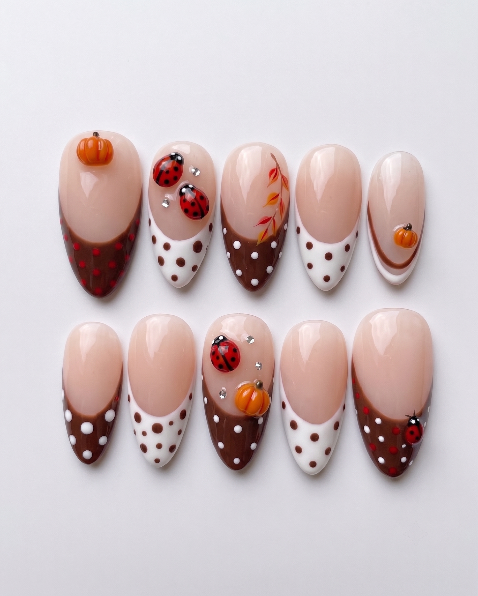 A set of press-on nails featuring brown French tips with white polka dots, decorated with 3D pumpkin charms and ladybug art.