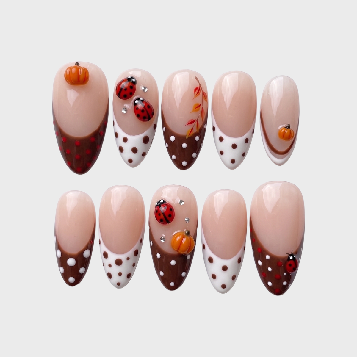 A set of press-on nails featuring brown French tips with white polka dots, decorated with 3D pumpkin charms and ladybug art.