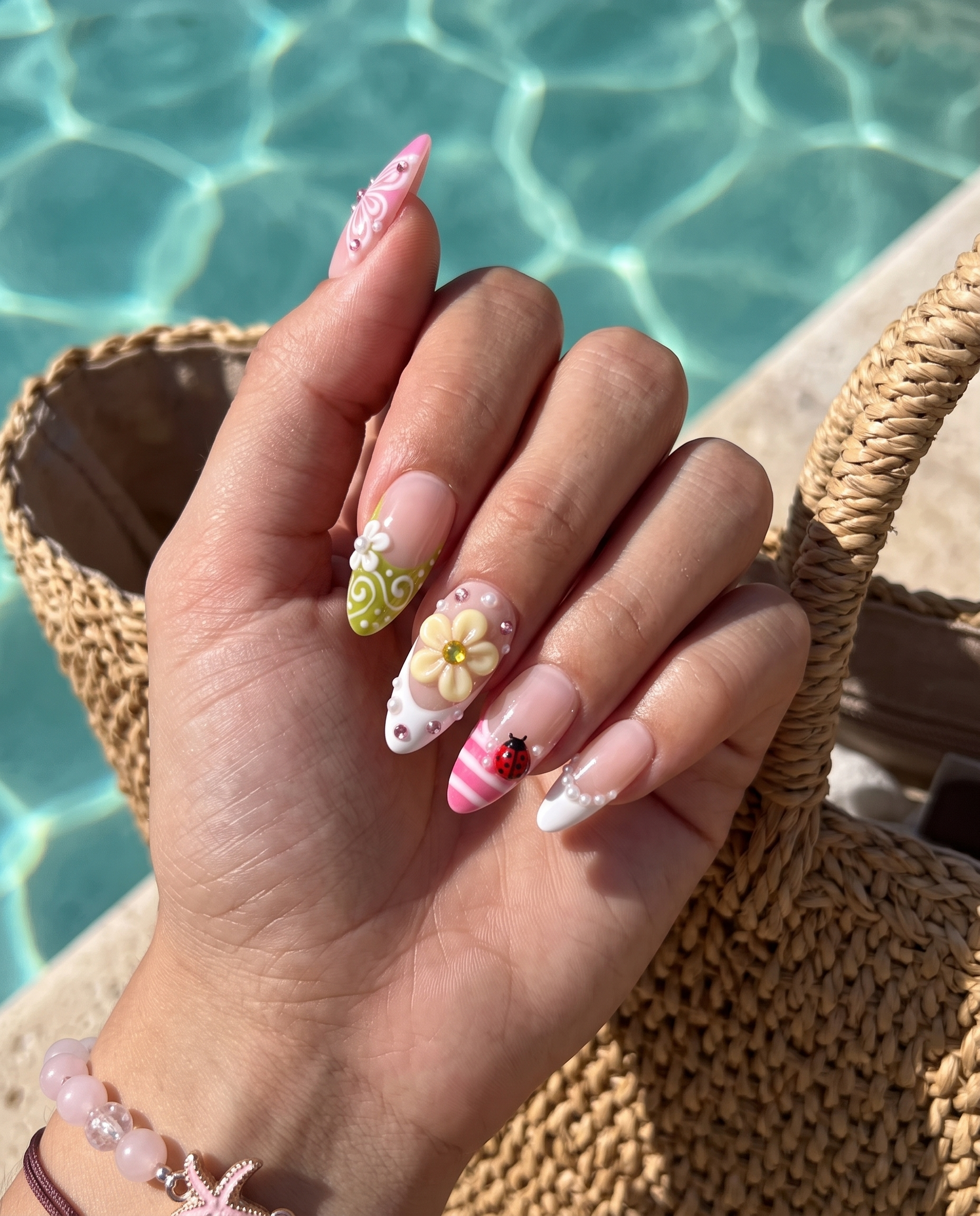 A set of glossy press-on nails decorated with 3D pink and yellow flowers, red ladybugs, white butterflies, and pearls on pink and green backgrounds.