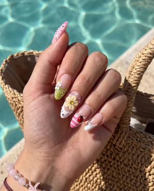A set of glossy press-on nails decorated with 3D pink and yellow flowers, red ladybugs, white butterflies, and pearls on pink and green backgrounds.
